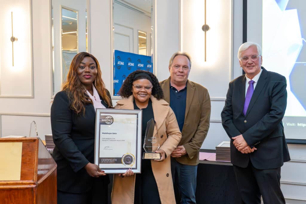 Four people pose at an awards ceremony; two women in front hold a framed certificate and a glass trophy, smiling with two men in the background.