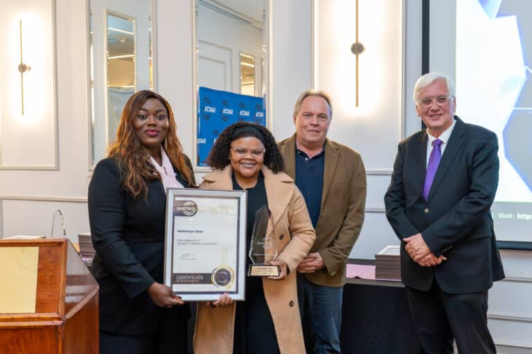 Four people pose at an awards ceremony; two women in front hold a framed certificate and a glass trophy, smiling with two men in the background.