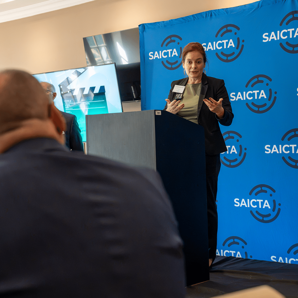 Woman speaker at a podium in front of a blue SAICTA backdrop, presenting to an audience.