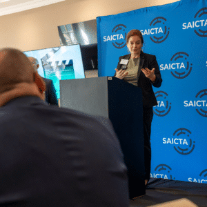 5 - SAICTA - SAICTA Woman speaker at a podium in front of a blue SAICTA backdrop, presenting to an audience.